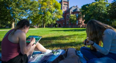 Two students sitting outside, one is drawing and the other is playing guitar